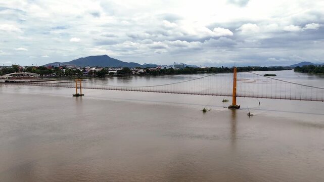 A wide aerial view of the Somphot Krung Rattanakosin 200 Pee suspension bridge crossing a wide, muddy river, with a town and mountain in the background under an overcast sky in Thailand.