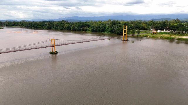 An aerial drone sequence of the Somphot Krung Rattanakosin 200 Pee suspension bridge spanning a wide muddy river, showcasing a lush green riverbank, distant mountains, and warm sunset reflections on t