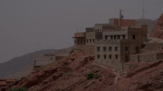 Multi-story concrete and earthen buildings cling to the jagged red rocky cliffside of a mountain village, depicting ongoing vernacular construction and rural resilience in Morocco&rsquo;s High Atlas.