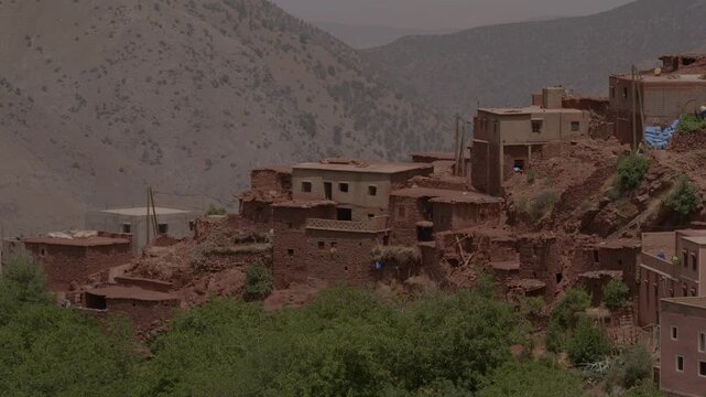 A cluster of red clay and stone Berber dwellings clings to a steep mountain ridge above a lush river valley oasis, capturing iconic vernacular architecture in Morocco&rsquo;s High Atlas Mountains region.
