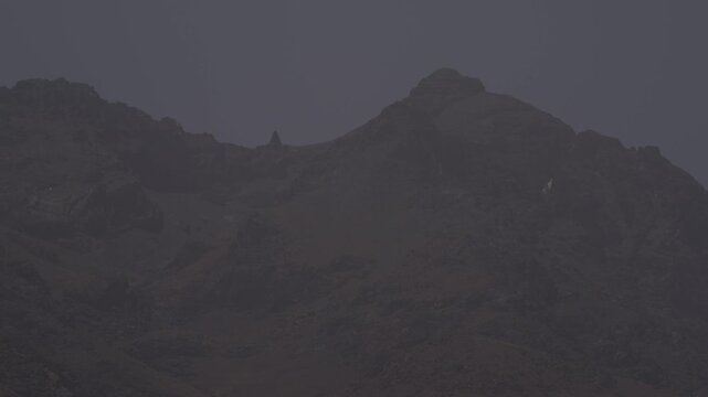 A rugged, barren mountain peak looms ominously under dark, heavy overcast skies, capturing the raw, brooding wilderness and dramatic high-altitude atmosphere of Morocco&rsquo;s High Atlas Mountains.