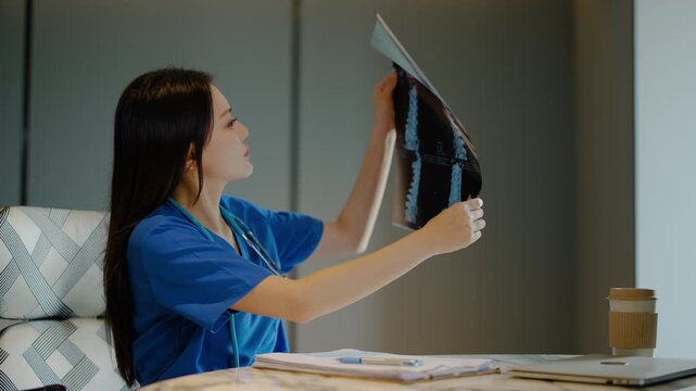 Asian female doctor in blue scrubs analyzing spine x-ray film at her desk in a clinic office, making medical diagnosis and writing patient notes on a clipboard, healthcare professional at work.