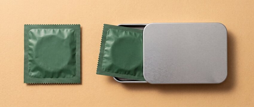 Green condom wrappers placed next to a metal tin container on a tan background with resting inside rectangular surface one wrapper two green square contraceptive reproductive preventative health