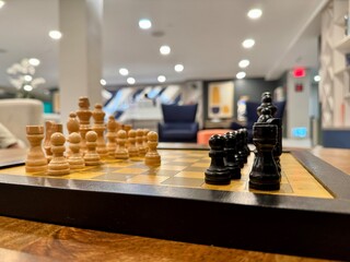 Close-up view of a chessboard with black and natural wood pieces arranged for play on a table in a modern indoor lounge, photographed from a low side angle with shallow depth of field. © Solomon Thompson