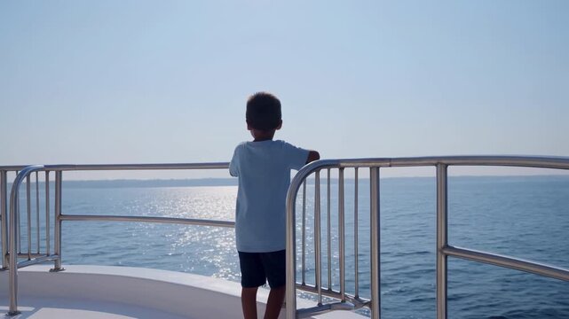 Child remains leaning on polished metal railing of a boat deck while looking across sparkling blue water toward a distant shoreline, seen from behind beneath clear bright sky and sunlight