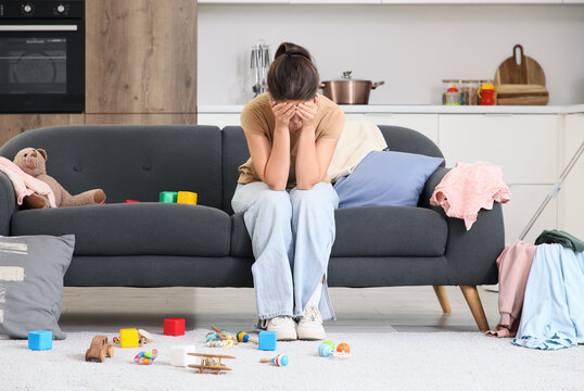 Parental burnout and mental health struggle, exhausted mother sitting on sofa in messy living room with scattered toys and household chores