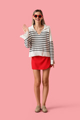 Friendly greeting and positive communication, happy young woman waving hand wearing stylish striped sweater and red skirt on a solid pink background © Pixel-Shot
