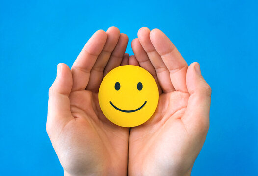 Hands holding cheerful yellow smiley face symbol against blue sky background