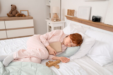 Fototapeta premium Cute teenage happy redhead girl in pajamas with adventure book and wooden airplane sleeping at home