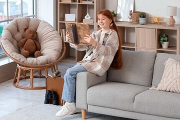 Fototapeta premium Cute teenage happy redhead girl with adventure book and suitcase sitting on sofa at home