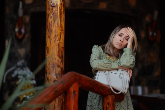 Tired Woman Feeling Sad While Standing On Wooden Veranda. Exhausted female looks down while leaning against a porch railing with handbag 
