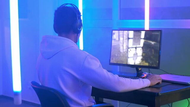 A young man wearing a light hoodie plays a first-person shooter game on his computer at a neon-lit indoor gaming station during nighttime.