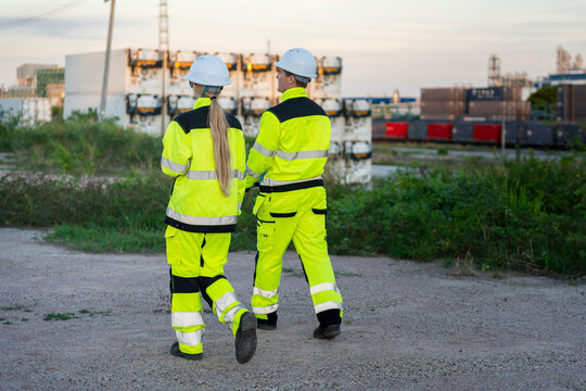 Male and female engineers in high visibility safety gear walking together at industrial plant yard, industrial technicians walking outdoors at shipping terminal facility during work inspection