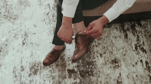Man in dark trousers sits on a stone wall and ties the laces of his brown leather brogues while getting ready for a wedding.