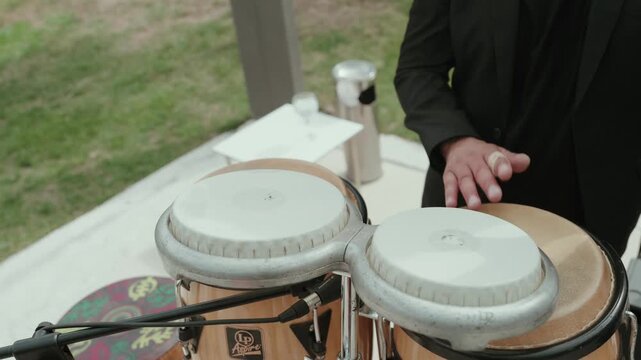 Hands of a percussionist play bongo drums beside a cymbal at a live music performance outdoors during a wedding party.