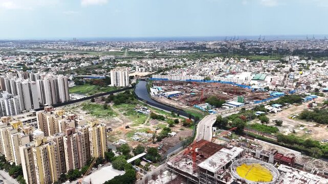 Wide aerial panorama of Chennai city, featuring modern high-rise residential towers, a canal or waterway, connecting roads and bridges, and a large-scale construction site with cranes.