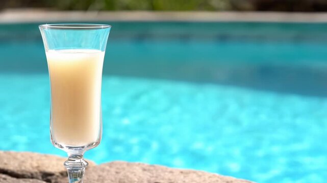Tall clear stemmed glass with pale creamy beverage rests on sunlit stone beside turquoise swimming pool as rippling water shimmers across softly blurred outdoor background