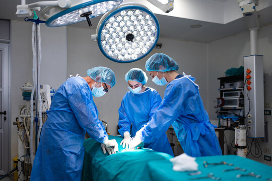Professional medical team performing surgery in a modern hospital operation room. Skilled doctors and surgeons wearing sterile blue gowns and masks focused on a surgical procedure under large lights.