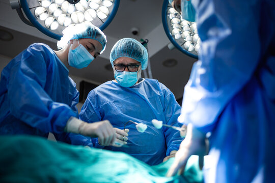 Professional medical doctors performing a surgical procedure in a bright operating room. A team of skilled surgeons wearing blue gowns masks and caps operate on a patient under bright surgical lights.