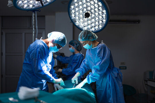 Dedicated medical doctors performing an operation in a dark hospital operating room. Expert surgeons wearing blue surgical gowns and face masks focused on a surgical procedure under bright lights.