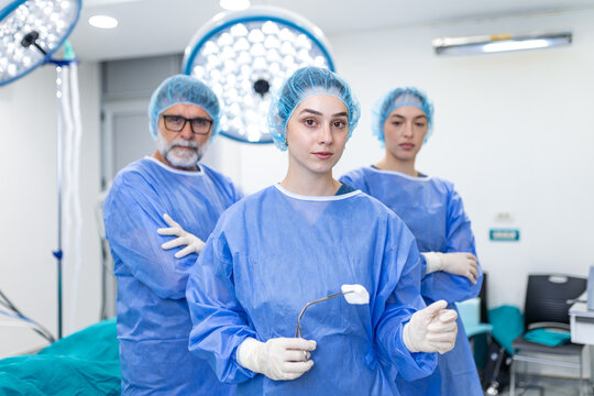 Team of medical doctors standing in an operating theatre room during surgery. Professional surgical staff wearing sterile blue gowns, face masks, and caps looking directly into camera together.