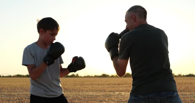 Father and son practice boxing together in sunlit field. Dad and teen spar with gloves during golden hour. Outdoor training builds fitness, confidence, and family teamwork. Fitness and family bonding.