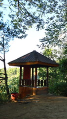 A rustic wooden gazebo with a dark roof stands amidst lush green trees and foliage under a bright sky, offering a serene view of the natural landscape. © Luthfia