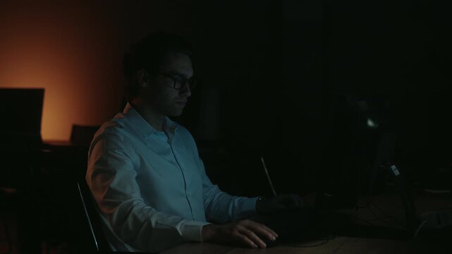 Ambitious young professional designer with glasses sitting at desk in dark room, working late at night and drawing on graphics tablet with pen, focused on creative project.