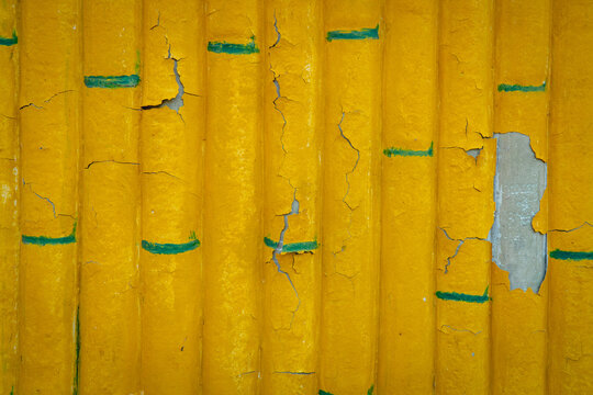 Yellow painted bamboo surface with peeling paint, cracks, and exposed blue-green layers creating an abstract weathered texture.