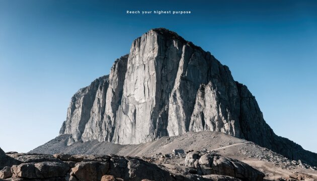 Isolated massive granite mountain peak with steep vertical rock faces and a jagged summit under a clear blue sky, showing a gravel path and rocky terrain at the base in daylight.