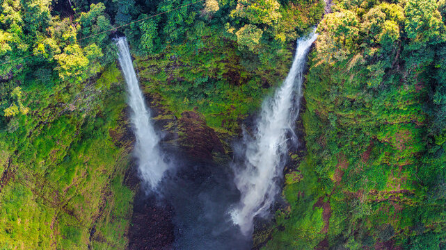 Aerial view of Tad Fane Waterfall in Laos, with a tourist enjoying tea while suspended on a zipline above the spectacular twin cascades and lush rainforest of the Bolaven Plateau.