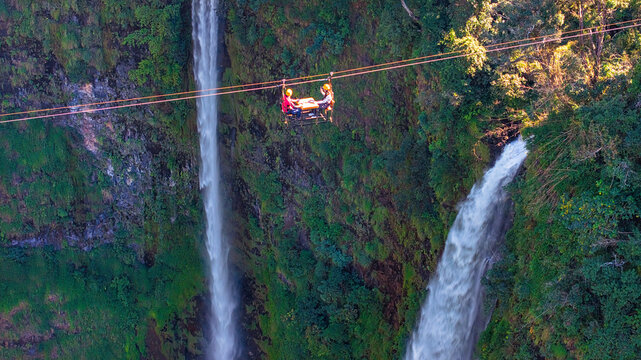 Aerial view of Tad Fane Waterfall in Laos, with a tourist enjoying tea while suspended on a zipline above the spectacular twin cascades and lush rainforest of the Bolaven Plateau.