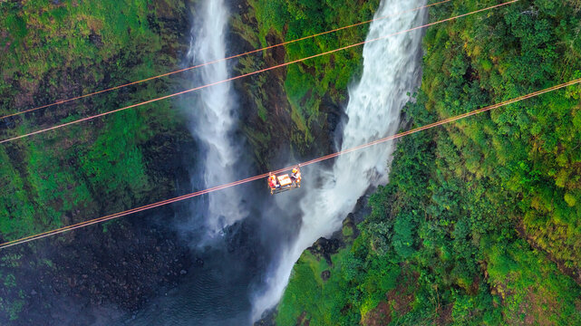 Aerial view of Tad Fane Waterfall in Laos, with a tourist enjoying tea while suspended on a zipline above the spectacular twin cascades and lush rainforest of the Bolaven Plateau.
