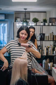 Female hairstylist with black hair using a hairdryer on a client with long blonde hair in a modern salon with shelves of hair products in the background
