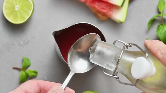 Clear sugar syrup is measured into watermelon juice to sweeten homemade summer cocktail mixture.