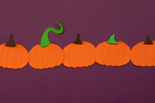 Paper pumpkins aligned in the center on a purple background, with decorative shapes and details, concept of fall crafts and decoration