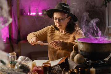 realistic witch brewing potion in cauldron. sitting at table making photo of spells on mobile phone. Everyday life intertwined with traditional witchcraft, magic and mystical atmosphere. © Andrii Lysenko