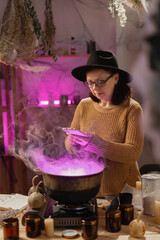 realistic witch brewing potion in cauldron. using mobile phone. Everyday life intertwined with traditional witchcraft, magic and mystical atmosphere. © Andrii Lysenko