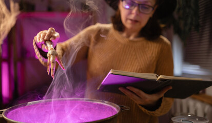 Halloween. Witch in hat witchcrafting, casting spells from spell book brews a magic potion in cauldron on fire in scary kitchen. Fantasy. © Andrii Lysenko