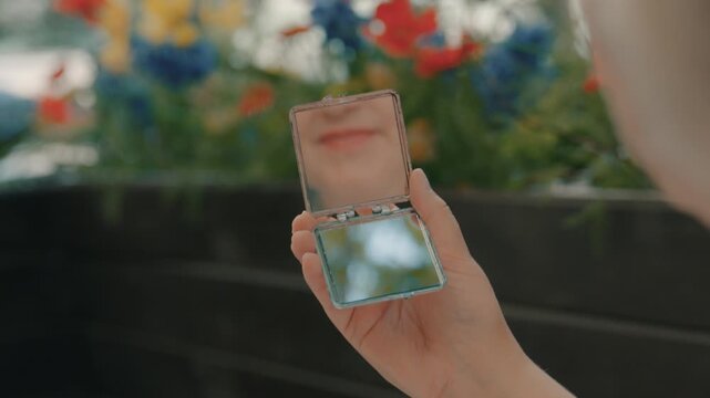 The woman&rsquo;s mouth is reflected in the pocket mirror she is holding in her hand while sitting on the street.