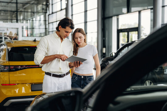 Male car sales representative in white shirt shows tablet to female customer in casual outfit inside modern car dealership with yellow vehicle in background