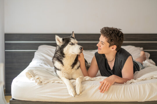 Teenager strokes back of husky resting beside him on white sheets, friendship stronger than many human ones, calm mood of home morning. Wide shot. Concept of friendship between teenager and dog