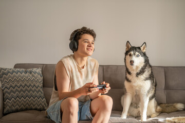 Fluffy husky sits beside teenage boy playing video game on soft sofa, hobby shared with patient waiting dog, hobby parents argue about. Wide shot. Concept of dog keeping company during game © iwavephoto