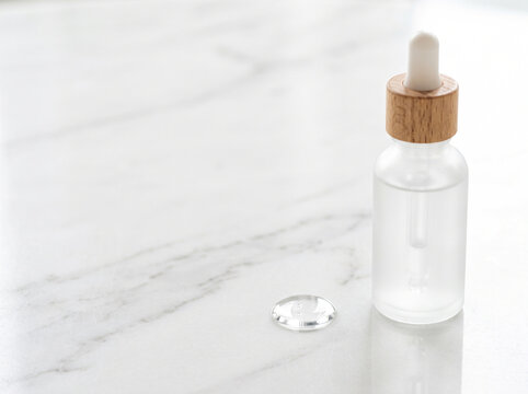 Glass serum dropper bottle with pipette and drop of liquid on white marble background with copy space for skincare wellness cosmetics concept