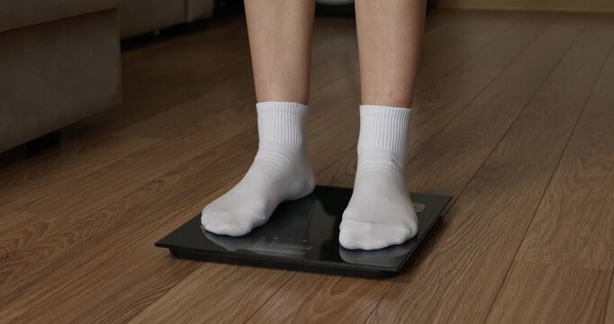 Woman feet in white socks stepping on digital electronic scales to check weight on wooden floor.