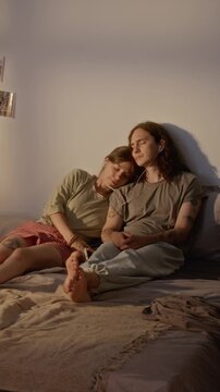 Vertical shot of young Caucasian couple sitting on bed sharing wired earphones while relaxing with closed eyes. Footage fits music streaming campaigns, wellness apps, and youth lifestyle marketing