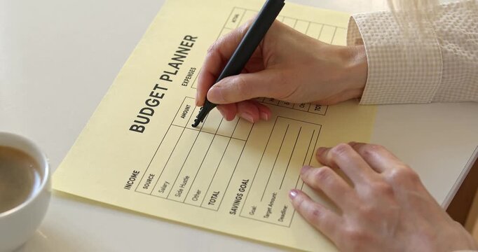 Woman hand writing income numbers with marker on paper budget planner next to coffee cup.