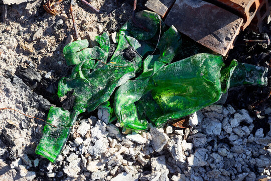 Deformed green glass bottles melted in a fire heap among gray ash and coal, creating an abstract texture of ruined trash after a campfire, high-angle close-up view outdoors