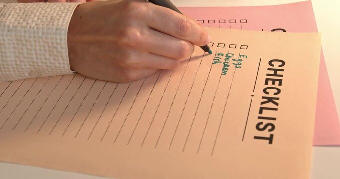 Close up of female hand writing on grocery checklist, filling food list with green marker.