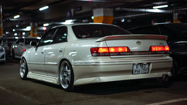 Tuning white Toyota Mark II X100 sedan car parked at underground parking lot. Rear view of classic Japanese JDM custom vehicle with alloy wheels, spoiler, taillights on and stance lifestyle concept.
